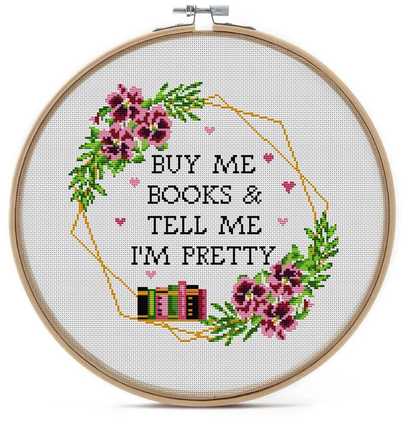 A bookish cross-stitch pattern with the text "BUY ME BOOKS & TELL ME I'M PRETTY". The design includes a floral wreath with pink flowers, green leaves, and a gold geometric shape. Small pink hearts and stacks of books are also included.
