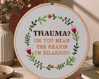 Puede incluir: Un bastidor de bordado circular de madera con tela Aida blanca y un diseño de punto de cruz. El texto "TRAUMA? OH YOU MEAN THE REASON I'M HILARIOUS" aparece en marrón oscuro y naranja, rodeado por una corona floral de hojas verdes y flores.