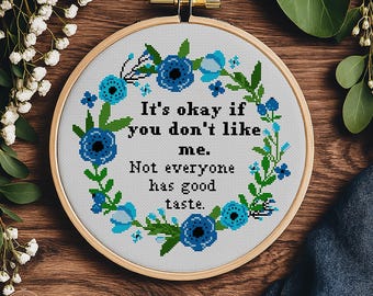 May include: A cross-stitch embroidery hoop with the text "It's okay if you don't like me. Not everyone has good taste." The design features blue floral accents and is set against a white background. The words "CROSS-STITCH PDF PATTERN" are at the bottom.