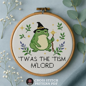 May include: Cross-stitch pattern featuring a green frog wearing a witch hat and holding a staff. The text reads "'TWAS THE 'TISM M'LORD" in black letters. The design is framed in a wooden hoop, with floral accents.
