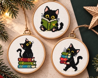 May include: Three circular wooden embroidery hoops containing cross-stitch patterns of a black cat with yellow eyes. One cat reads a green book, one sits on a stack of red, green, and blue books, and one carries a stack of books. Text reads CROSS STITCH PATTERNS PDF.