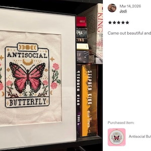 K&ouml;nnte beinhalten: Gerahmtes Kreuzstich-Kunstwerk mit dem Text "ANTISOCIAL BUTTERFLY" und einem Schmetterlingsdesign. Der Schmetterling ist rosa und schwarz, umgeben von rosa Rosen und gr&uuml;nen Bl&auml;ttern. Das Kunstwerk befindet sich in einem wei&szlig;en Rahmen.