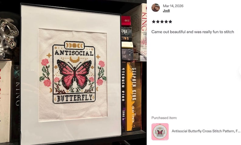 K&ouml;nnte beinhalten: Gerahmtes Kreuzstich-Kunstwerk mit dem Text "ANTISOCIAL BUTTERFLY" und einem Schmetterlingsdesign. Der Schmetterling ist rosa und schwarz, umgeben von rosa Rosen und gr&uuml;nen Bl&auml;ttern. Das Kunstwerk befindet sich in einem wei&szlig;en Rahmen.