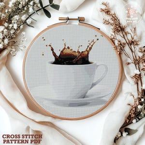 Op de afbeelding: Een kruissteekpatroon in een houten borduurring met een witte koffiekop op een bijpassende schotel waar donkerbruine koffie uit spat. De stoffen achtergrond is lichtgrijs en de tekst CROSS STITCH WITH ART is zichtbaar op de afbeelding.