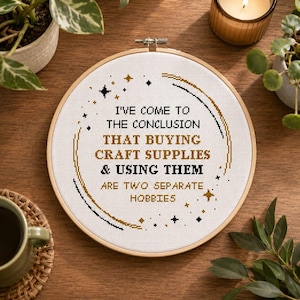 May include: A circular wooden embroidery hoop containing white fabric with black and gold cross-stitched text that reads: I'VE COME TO THE CONCLUSION THAT BUYING CRAFT SUPPLIES & USING THEM ARE TWO SEPARATE HOBBIES, surrounded by small star motifs.