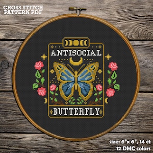 May include: A cross stitch pattern featuring a butterfly with blue wings and the text "ANTISOCIAL BUTTERFLY". The pattern is framed in a gold-colored border with a crescent moon and stars. The background is black and the pattern is 6 inches by 6 inches.