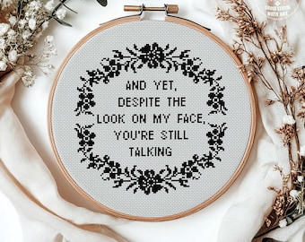 May include: A cross-stitch embroidery hoop with the text "AND YET, DESPITE THE LOOK ON MY FACE, YOU'RE STILL TALKING" in black. The text is surrounded by a black floral wreath. The words "CROSS STITCH PATTERN PDF" are at the bottom.