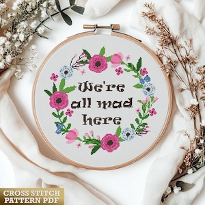 以下が含まれることがあります： 木製のフレームに入ったクロスステッチパターンで、「We're all mad here」の文字が花のリースで囲まれています。 リースにはピンクと青の花と緑の葉が含まれています。 下部には「CROSS STITCH PATTERN PDF」の文字があります。