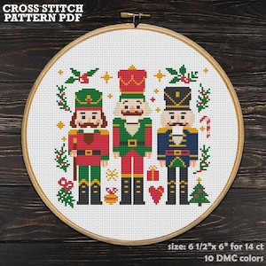 May include: A cross-stitch pattern featuring three toy soldiers in festive colors, set within a wooden embroidery hoop. The design includes Christmas elements like a tree, candy cane, and holly. The text "CROSS STITCH PATTERN PDF" is at the top. The size is 6.5" x 6" for 14 ct.