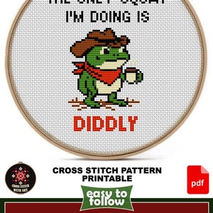 May include: Cross-stitch pattern in a wooden hoop. The design features a green frog wearing a cowboy hat and holding a coffee cup, with the text "THE ONLY SQUAT I'M DOING IS DIDDLY". The design size is 4 1/2" x 5 1/2".