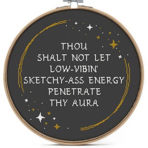 May include: A circular wooden embroidery hoop containing black fabric with white cross-stitched text that reads: THOU SHALT NOT LET LOW-VIBIN' SKETCHY-ASS ENERGY PENETRATE THY AURA. The design includes gold and white stitched stars and a curved gold line.