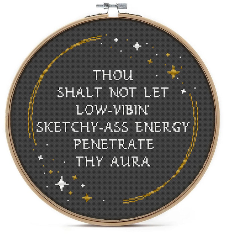 May include: A circular wooden embroidery hoop containing black fabric with white cross-stitched text that reads: THOU SHALT NOT LET LOW-VIBIN' SKETCHY-ASS ENERGY PENETRATE THY AURA. The design includes gold and white stitched stars and a curved gold line.