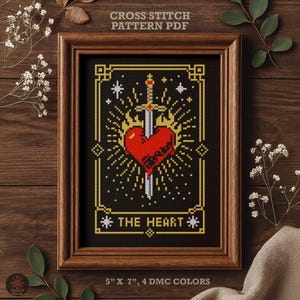 May include: Framed cross-stitch pattern featuring a red heart pierced by a sword, with the text "THE HEART" below. The design is in gold and white on a black background, framed in a wooden frame. The text "CROSS STITCH PATTERN PDF" is at the top. The dimensions are 5" x 7".