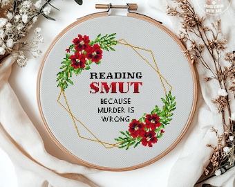 May include: A cross-stitch embroidery hoop with the text "Reading Smut Because Murder Is Wrong." The design features red flowers and green leaves, with a gold geometric outline. The wooden hoop is set against a white background.
