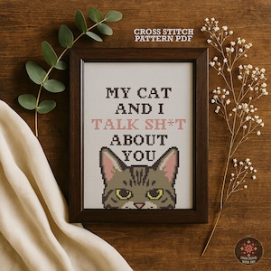 Może przedstawiać: Oprawiony wzór haftu krzyżykowego z napisem "MY CAT AND I TALK SH*T ABOUT YOU" i twarzą kota. Rama jest ciemnobrązowa, a tło białe. Obraz jest ozdobiony gałązkami zieleni i kwiatów.