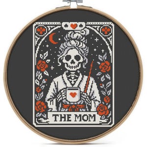 A cross-stitch design in a wooden hoop features a skeleton figure holding a mug with a heart. The design includes red roses, a crescent moon, and the text "THE MOM" on a banner. The background is black.