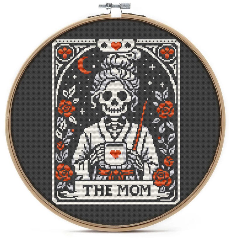 A cross-stitch design in a wooden hoop features a skeleton figure holding a mug with a heart. The design includes red roses, a crescent moon, and the text "THE MOM" on a banner. The background is black.