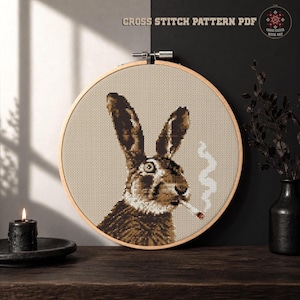 May include: A cross stitch pattern PDF featuring a brown hare with long ears, holding a lit cigarette with white smoke rising, displayed in a round wooden embroidery hoop. The text CROSS STITCH PATTERN PDF is visible at the top of the image.