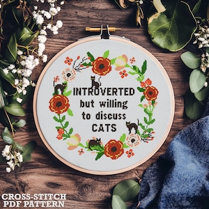 Könnte beinhalten: Stickmuster mit einem Kranz aus roten und rosa Blumen mit schwarzen Katzen und dem Text "Introvertiert, aber bereit, über Katzen zu diskutieren".
