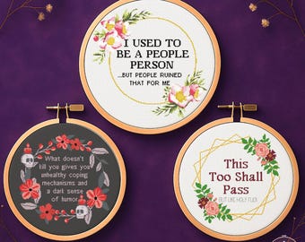 May include: Three cross-stitch patterns in wooden hoops. The top one reads "I USED TO BE A PEOPLE PERSON...BUT PEOPLE RUINED THAT FOR ME." The bottom left reads "What doesn't kill you gives you unhealthy coping mechanisms and a dark sense of humour."