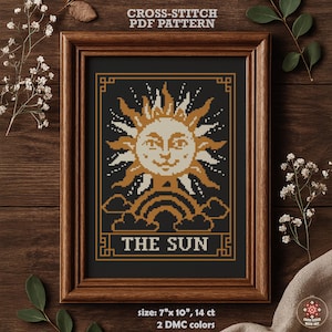 May include: A framed cross-stitch pattern featuring a smiling sun design with a rainbow and clouds. The text "THE SUN" is at the bottom. The frame is brown, and the pattern size is 7" x 10".