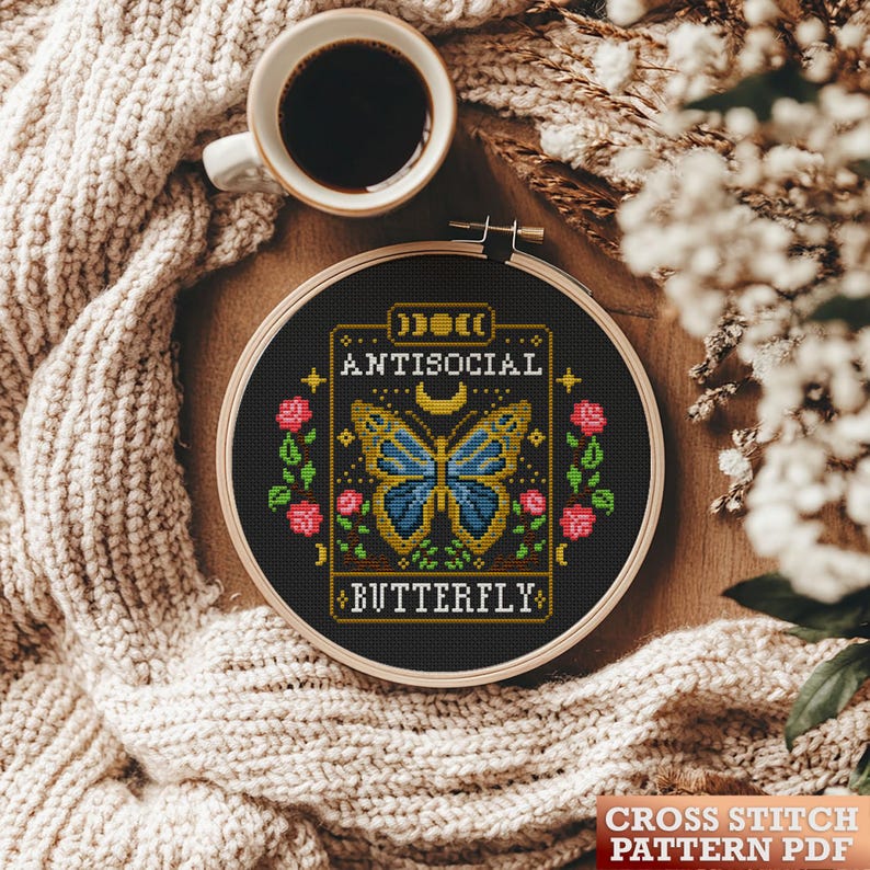 May include: A black and white cross stitch pattern featuring a butterfly with blue wings and a gold outline. The butterfly is surrounded by flowers and the text "Antisocial Butterfly" in a gold frame.