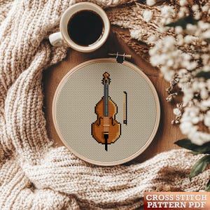 May include: A cross stitch pattern for a brown double bass with a bow. The pattern is displayed in a wooden embroidery hoop on a wooden table with a beige knitted sweater and white flowers.