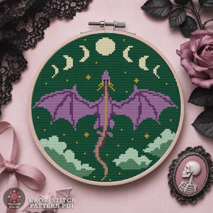 Puede incluir: Un bastidor de bordado circular de madera que contiene un diseño de punto de cruz de un dragón morado con las alas extendidas sobre un fondo verde oscuro. El diseño presenta un ciclo de fases lunares, estrellas amarillas y nubes verde claro.