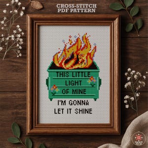 May include: A framed cross-stitch pattern showing a green skip on fire. The text "THIS LITTLE LIGHT OF MINE I'M GONNA LET IT SHINE" is stitched below. The frame is brown and the background is white. The words "CROSS-STITCH PDF PATTERN" are at the top.