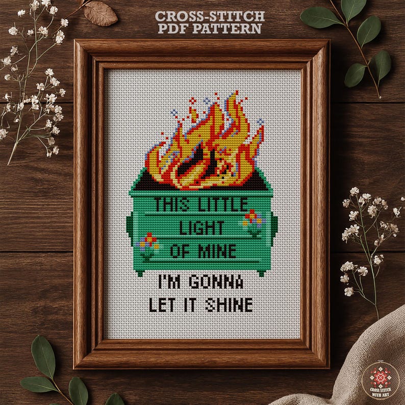 A framed cross-stitch pattern with a green dumpster on fire and text reads "THIS LITTLE LIGHT OF MINE, I'M GONNA LET IT SHINE." The pattern is 5" x 7" and uses 7 DMC colors, PDF download