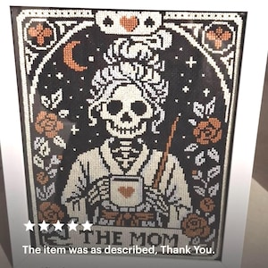 K&ouml;nnte beinhalten: Gerahmtes Kreuzstich-Kunstwerk mit einer Skelettfigur, die eine Tasse mit einem Herzen h&auml;lt. Das Design umfasst Rosen, einen Halbmond und den Text "THE MOM" in einem wei&szlig;en Rahmen. Die Farbpalette umfasst Schwarz, Wei&szlig; und Orange.