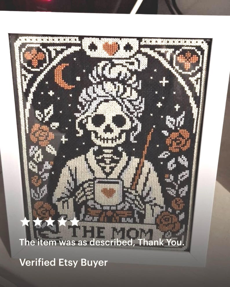 K&ouml;nnte beinhalten: Gerahmtes Kreuzstich-Kunstwerk mit einer Skelettfigur, die eine Tasse mit einem Herzen h&auml;lt. Das Design umfasst Rosen, einen Halbmond und den Text "THE MOM" in einem wei&szlig;en Rahmen. Die Farbpalette umfasst Schwarz, Wei&szlig; und Orange.