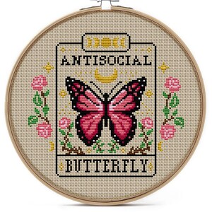 K&ouml;nnte beinhalten: Kreuzstich-Stickerei in einem Holzreifen. Das Design zeigt einen rosa und schwarzen Schmetterling, die Worte "ANTISOCIAL BUTTERFLY" und florale Akzente. Der Hintergrund ist ein hellbeiger Stoff.