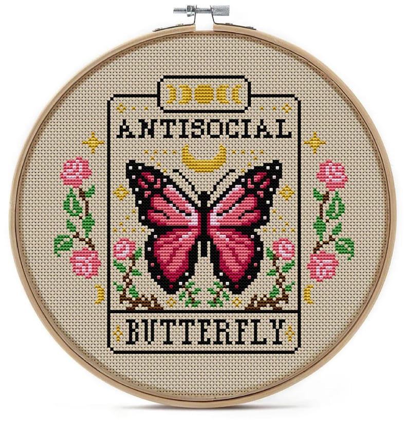 K&ouml;nnte beinhalten: Kreuzstich-Stickerei in einem Holzreifen. Das Design zeigt einen rosa und schwarzen Schmetterling, die Worte "ANTISOCIAL BUTTERFLY" und florale Akzente. Der Hintergrund ist ein hellbeiger Stoff.