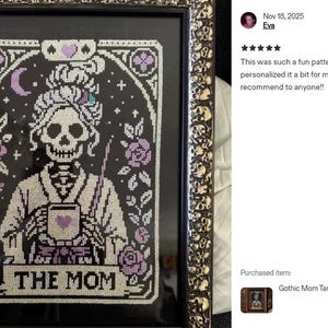 K&ouml;nnte beinhalten: Gerahmtes Kreuzstich-Kunstwerk mit einer Skelettfrau, die eine Tasse h&auml;lt, mit dem Text "THE MOM" darunter. Das Design umfasst lila Rosen und einen Mond auf schwarzem Hintergrund, in einem dekorativen Goldrahmen.