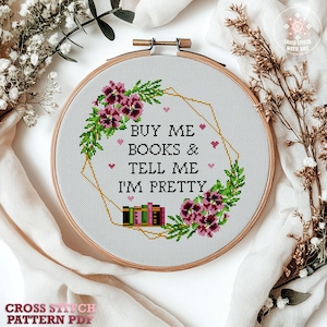 Cross-stitch embroidery in a wooden hoop. The design features the text "BUY ME BOOKS & TELL ME I'M PRETTY" surrounded by floral and geometric elements. The color palette includes pink, green, and gold.