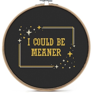 May include: A cross-stitch embroidery hoop featuring the phrase "I COULD BE MEANER" in yellow thread on a black fabric. The design is framed by a gold and white border with star motifs, set within a wooden hoop.