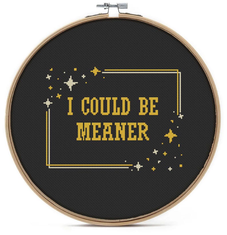 May include: A cross-stitch embroidery hoop featuring the phrase "I COULD BE MEANER" in yellow thread on a black fabric. The design is framed by a gold and white border with star motifs, set within a wooden hoop.