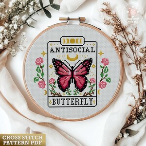 K&ouml;nnte beinhalten: Ein Kreuzstichmuster in einem Holzrahmen mit einem rosa Schmetterling und dem Schriftzug "Antisocial Butterfly". Das Design ist von rosa Rosen und Gr&uuml;n umgeben. Der Text "Cross Stitch Pattern PDF" befindet sich unten.