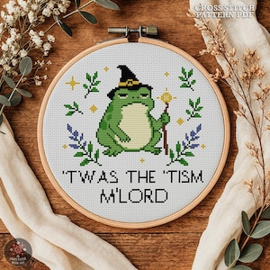 May include: A circular wooden embroidery hoop containing a cross stitch pattern of a green frog wearing a black witch hat and holding a staff. The text reads 'TWAS THE 'TISM M'LORD' in black letters, surrounded by small green leaves and yellow stars.