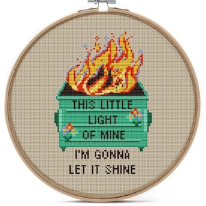 May include: Cross-stitch embroidery in a wooden hoop features a green dumpster engulfed in flames, with the text "THIS LITTLE LIGHT OF MINE I'M GONNA LET IT SHINE." The flames are yellow, orange, and red.