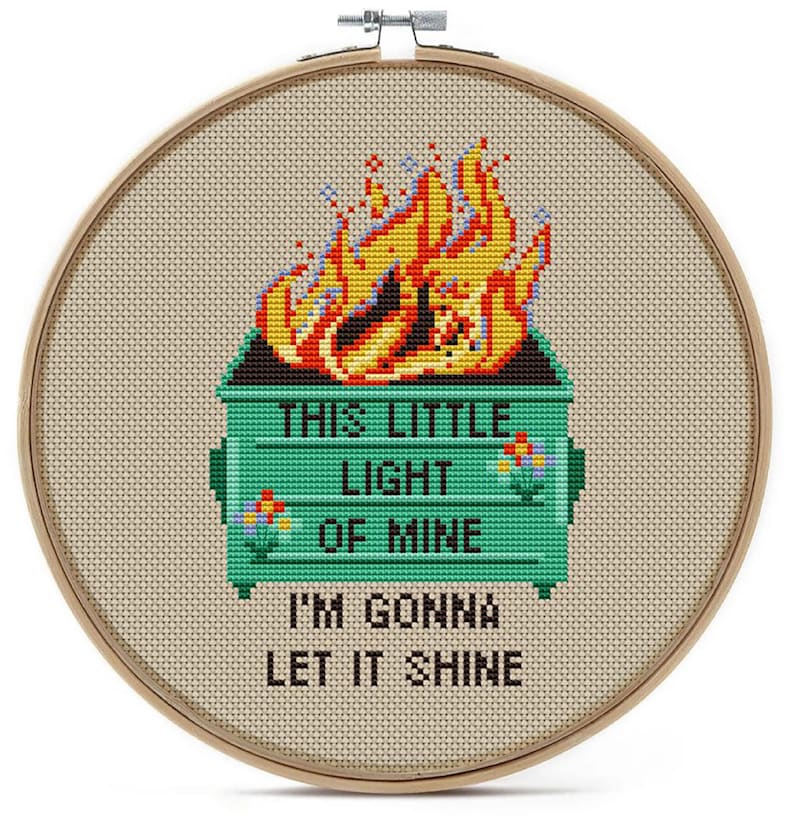 May include: Cross-stitch embroidery in a wooden hoop features a green dumpster engulfed in flames, with the text "THIS LITTLE LIGHT OF MINE I'M GONNA LET IT SHINE." The flames are yellow, orange, and red.