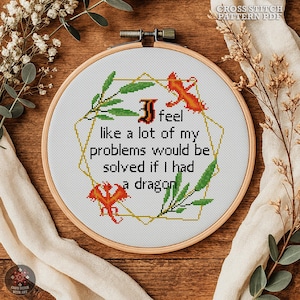May include: A cross-stitch embroidery hoop featuring the text "I feel like a lot of my problems would be solved if I had a dragon." The design includes orange dragons, green leaves, and a gold geometric border. The hoop is set against a wooden background.