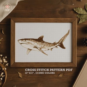 May include: A framed cross-stitch pattern of a tiger shark in shades of brown. The artwork is displayed in a wooden frame and set against a neutral background. The text "CROSS STITCH PATTERN PDF 12" X 6", 10 DMC COLORS" is visible.