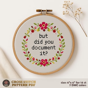 以下が含まれることがあります： 「but did you document it?」の文字が花輪で囲まれたクロスステッチ刺繍枠。 刺繍は木製の枠の中にあり、文字は黒、花はピンク、赤、緑です。