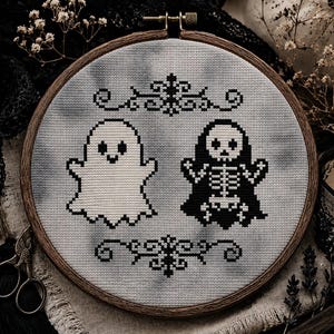 May include: A circular wooden embroidery hoop containing a cross-stitch design on grey fabric. The design features a white ghost and a black skeleton figure, both centered between two black decorative scrollwork patterns stitched in black thread.