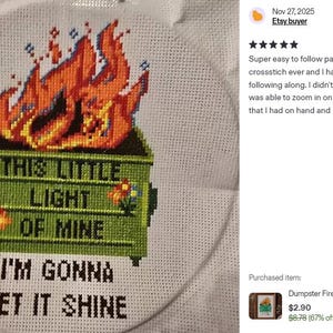 Cross-stitch embroidery on white fabric, featuring a green dumpster engulfed in orange flames. The text reads "THIS LITTLE LIGHT OF MINE, I'M GONNA LET IT SHINE." The design is framed in a circular hoop.
