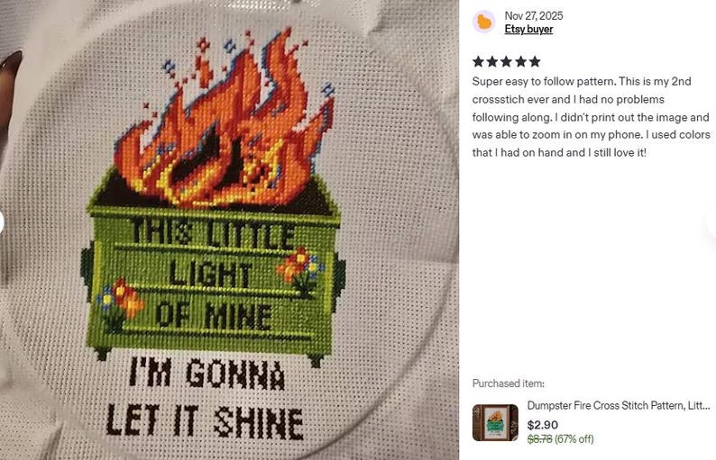 Cross-stitch embroidery on white fabric, featuring a green dumpster engulfed in orange flames. The text reads "THIS LITTLE LIGHT OF MINE, I'M GONNA LET IT SHINE." The design is framed in a circular hoop.