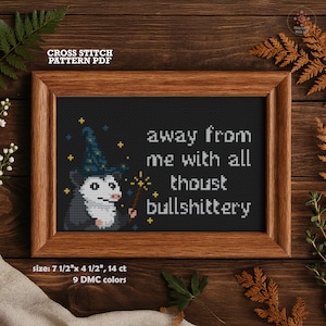 May include: Cross-stitch pattern PDF in a wooden frame. The design features a possum wearing a wizard hat and holding a wand, with the text "away from me with all thoust bullshittery." The framed piece measures 19 cm x 11.4 cm.