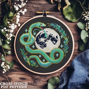 May include: A cross-stitch pattern featuring a green snake wrapped around a black and white moon with a floral design. The pattern is in a circular embroidery hoop.